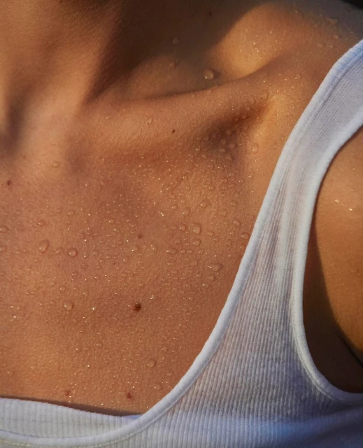 Close-up of a fair-skinned adult female's upper chest and shoulders glistening with water droplets, wearing a white tank top.