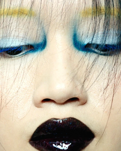 Close-up portrait of a person with bold blue and yellow eye makeup and dark lipstick, emphasizing beauty and fashion.