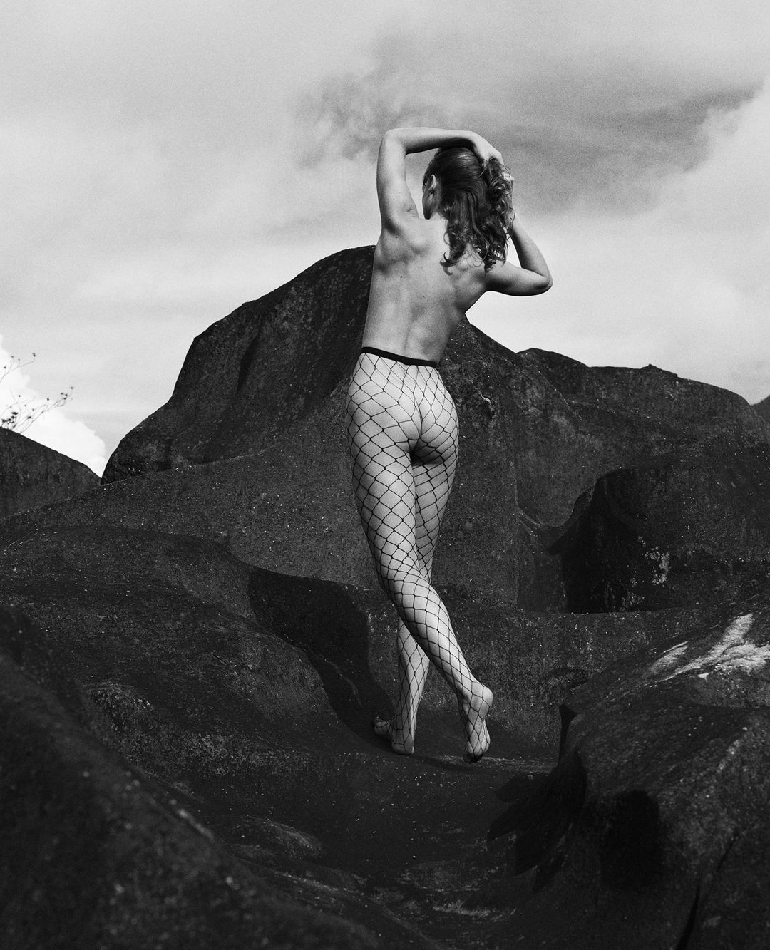 Monochrome photograph of a woman wearing fishnet tights posing amidst dark rocky terrain under an overcast sky.