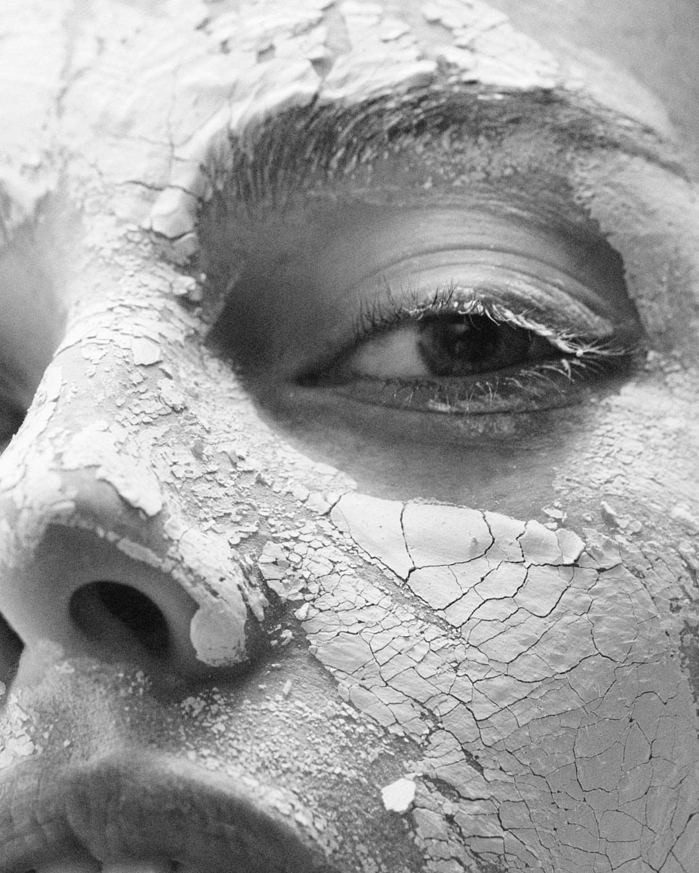 Close-up black and white shot of an adult female's face covered in cracked, peeling clay or mud mask, focused on her eye.