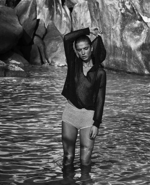 A woman in a hooded mesh top stands in shallow water with rocky cliffs in the background, creating a natural and serene fashion image.