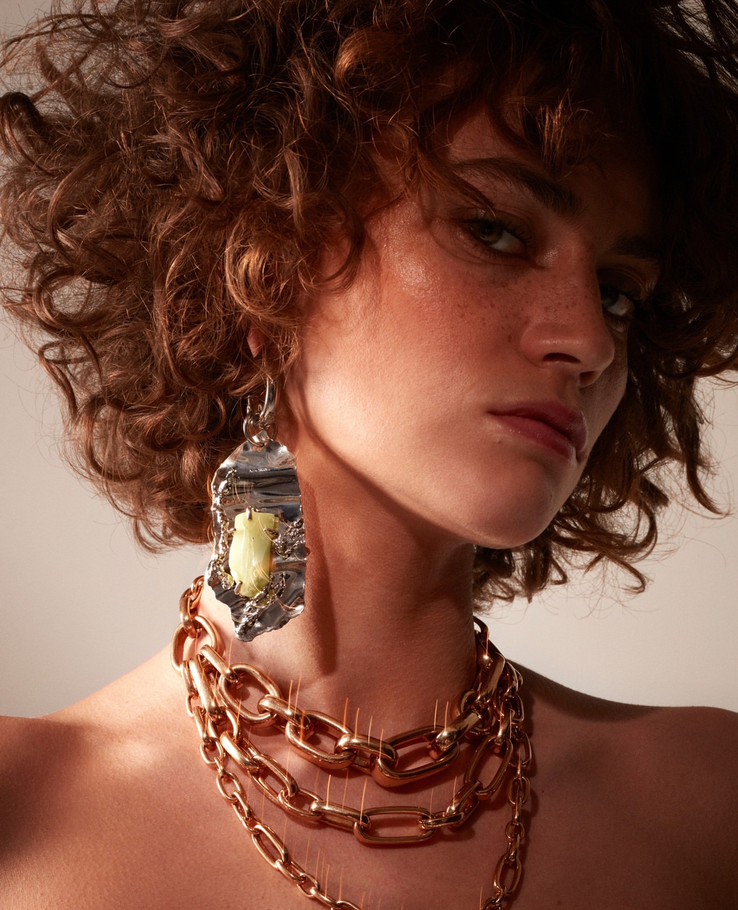 A woman with curly hair wearing statement jewelry featuring earrings with silver and gold tones and a layered gold chain necklace.