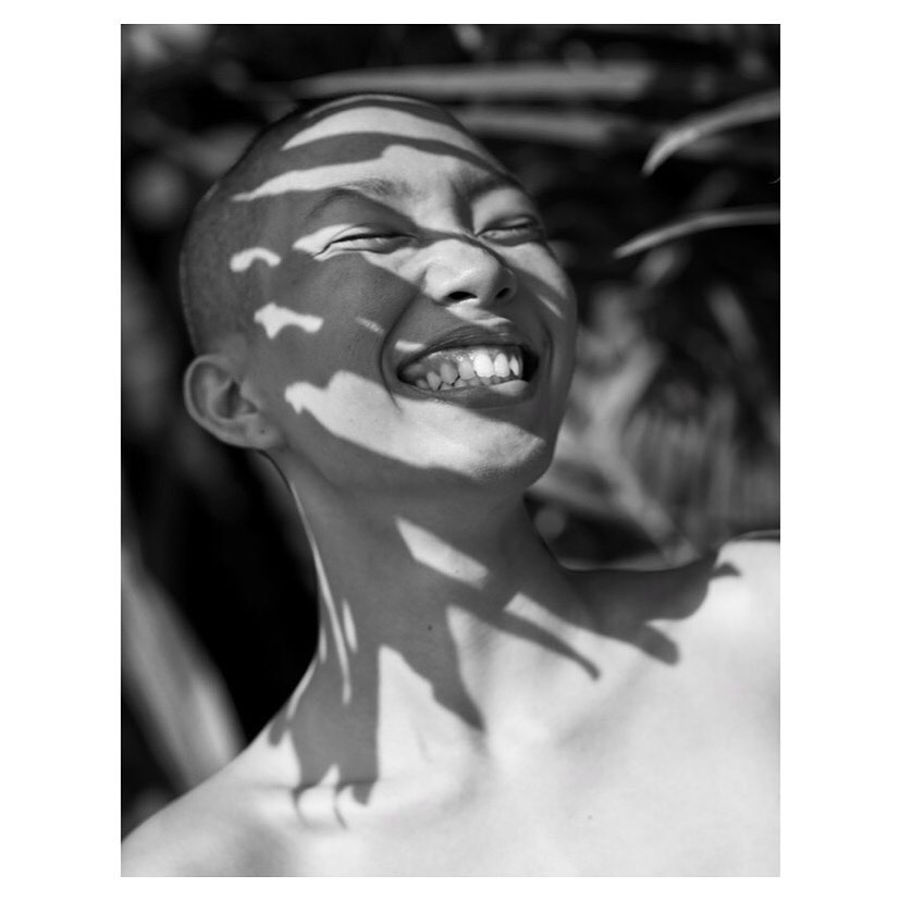 Black and white portrait of smiling person with a shaved head, sunlight dappling face with plant shadows, bright and cheerful.