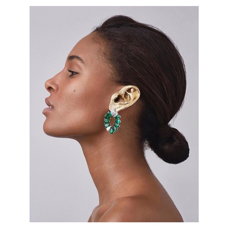 Profile shot of a dark-haired woman with olive skin wearing a decorative gold ear covering and green and white earrings on a grey background.