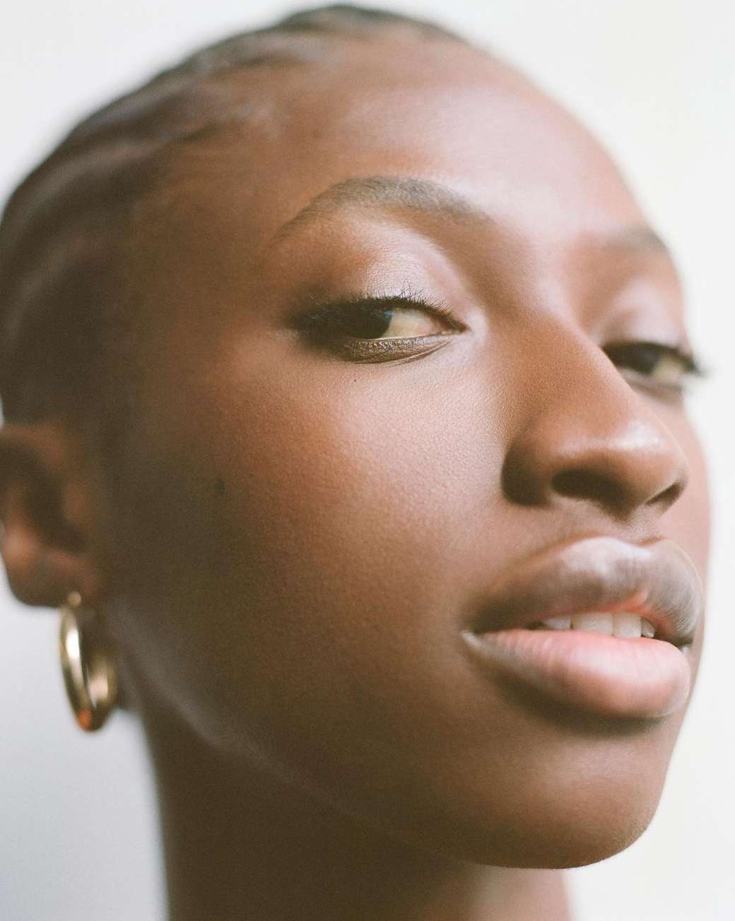 Close-up portrait of a young Black woman with gold hoop earrings against a neutral backdrop, showcasing beauty and minimalist style.