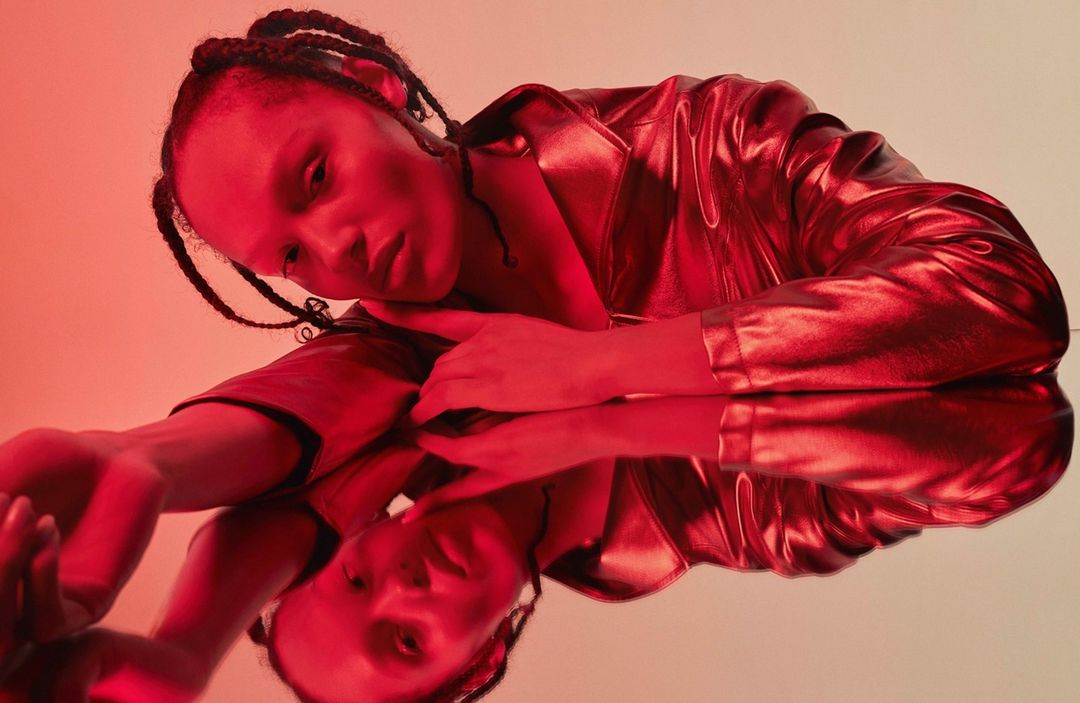 A young adult woman with braids rests her head on a reflective surface in a red-lit studio setting, creating a mirror image.