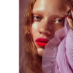 Close up portrait of a fair skinned woman with red hair wearing pink lipstick holding pleated lavender fabric to her face by Karina Twiss