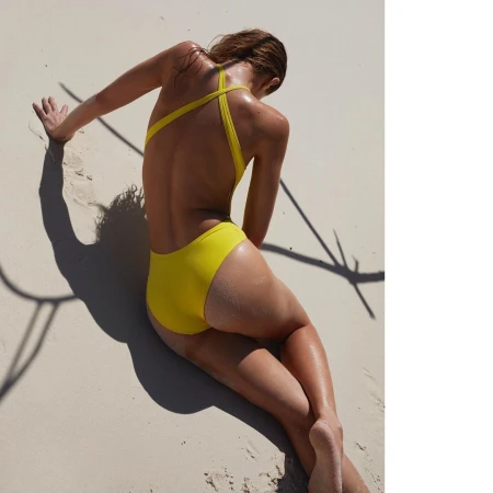Woman in a yellow one-piece swimsuit reclining on white sand beach under bright overhead sunlight casting deep shadows.