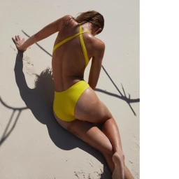 Woman in a yellow one-piece swimsuit reclining on white sand beach under bright overhead sunlight casting deep shadows.