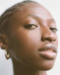 Close-up portrait of a Black woman with braided hair wearing gold hoop earrings against a neutral background.