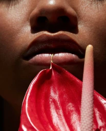 Close-up of a person's lower face holding a vibrant red anthurium flower near their lips under harsh sunlight.
