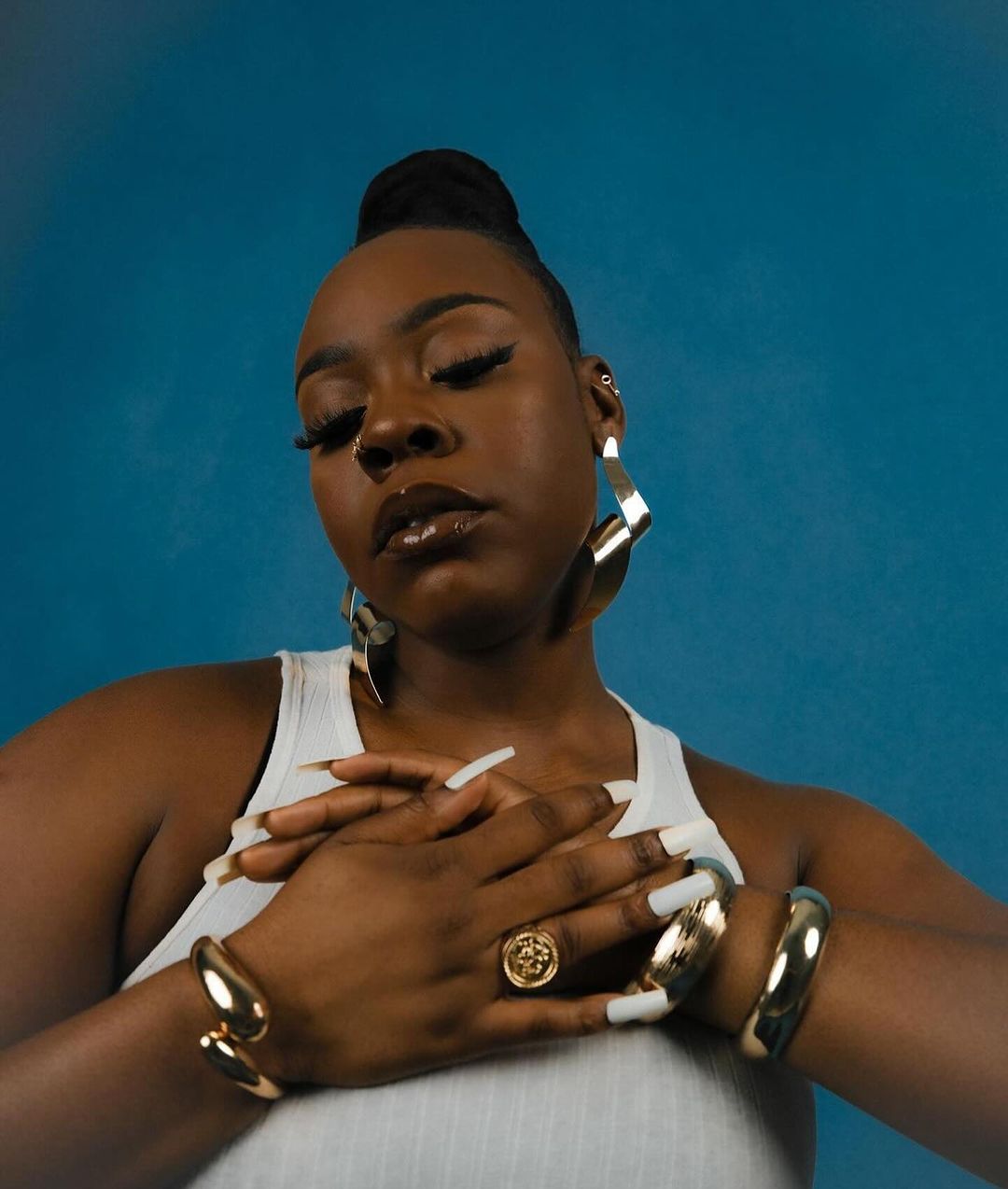 Close-up of a stylish Black woman with long nails posing against a blue backdrop, wearing gold jewelry, and a white tank top.