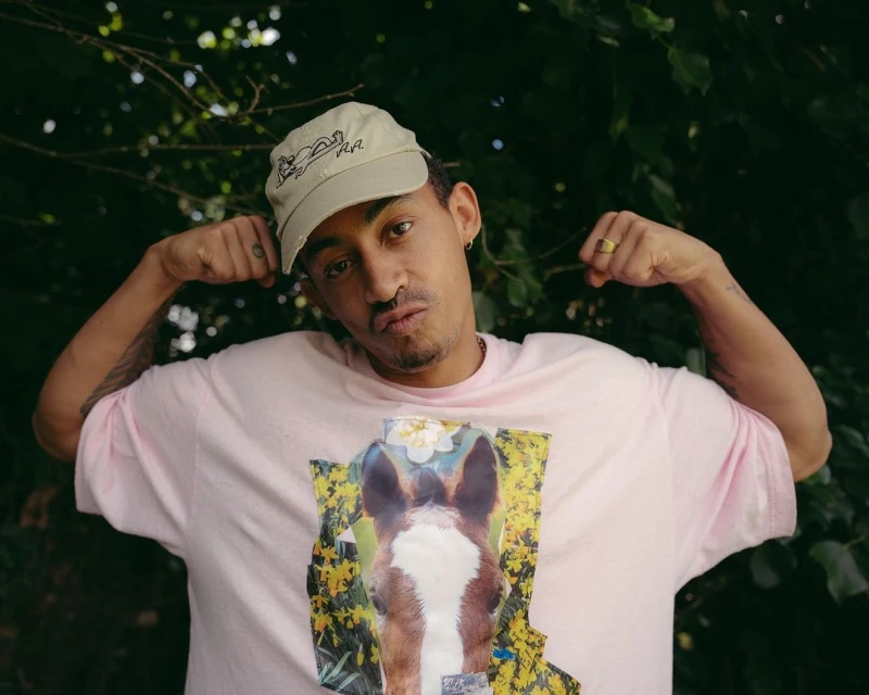 Adult male flexing biceps in a medium outdoor shot with leafy green foliage, wearing a light pink t-shirt and khaki hat.