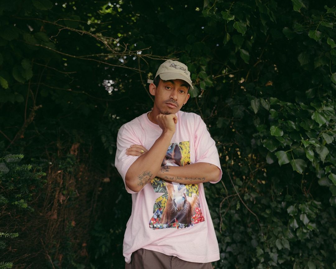 Portrait of a young adult male outdoors, wearing a printed t-shirt and a cap, with a leafy green background.