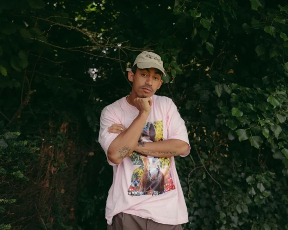 Portrait of a young adult male outdoors, wearing a printed t-shirt and a cap, with a leafy green background.