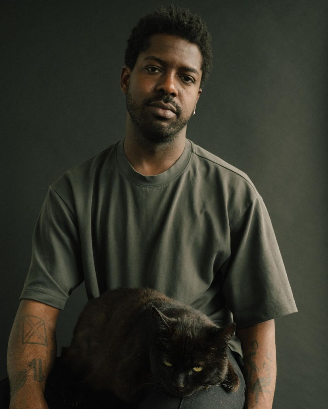 Portrait of adult Black male in t-shirt with Black cat sitting on lap against dark background. Tattoos visible on both arms.