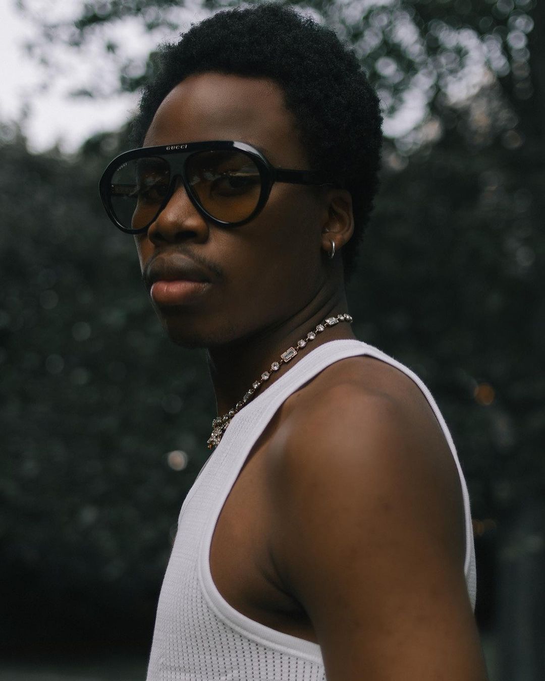 Portrait of an adult Black man wearing sunglasses and a white tank top outdoors against a blurred green leafy background.