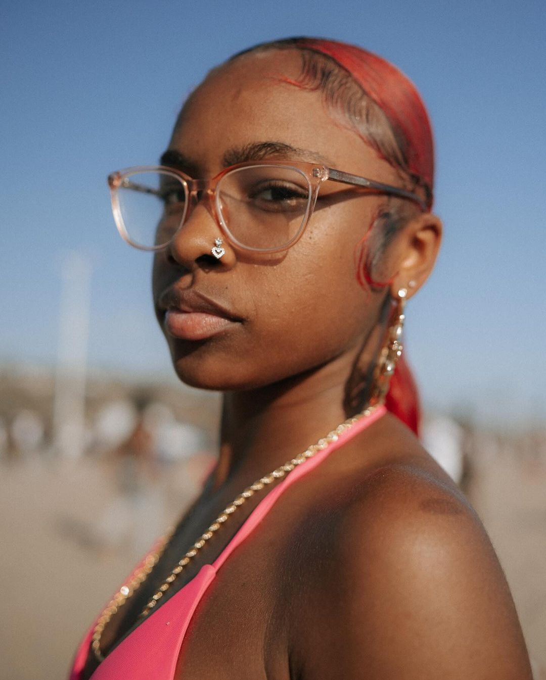Close-up portrait of a Black woman with red-tinted hair wearing glasses, a pink bikini top and a gold chain on a sunny day outdoors.