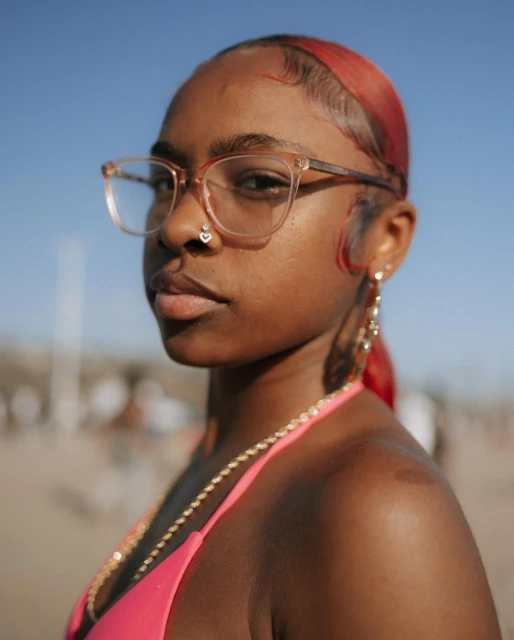 Close-up portrait of a Black woman with red-tinted hair wearing glasses, a pink bikini top and a gold chain on a sunny day outdoors.