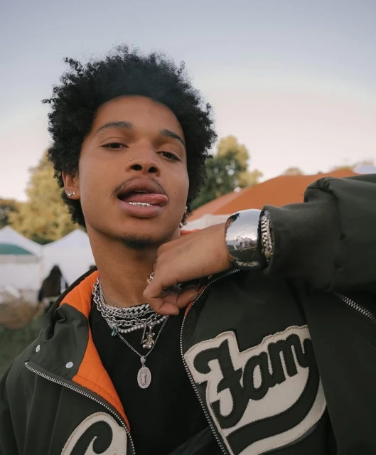 Portrait of a young man with dark curly hair wearing a jacket and jewelry, sticking out his tongue, in an outdoor setting.