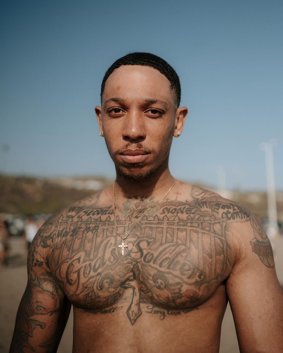 Portrait of a tatted, muscular man with a gold chain standing on a beach on a sunny day with the ocean and sky in the background.