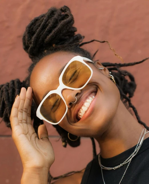 Close-up portrait of a smiling African-American woman with dreadlocks and white-framed sunglasses against a terracotta wall backdrop.