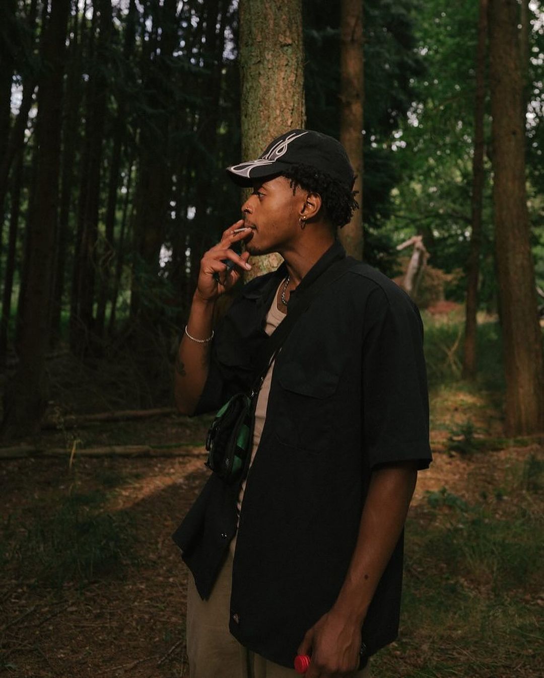 Young man smoking in a forest, wearing a baseball cap and black jacket, khaki pants, with tall trees in soft, natural light.