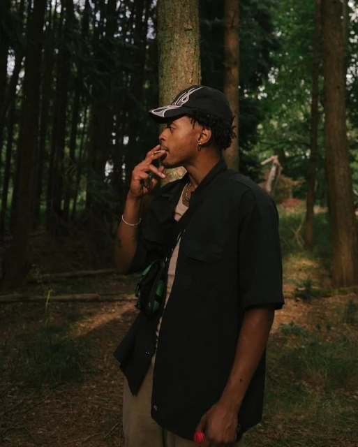 Young man smoking in a forest, wearing a baseball cap and black jacket, khaki pants, with tall trees in soft, natural light.