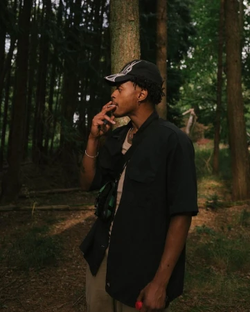 Young man smoking in a forest, wearing a baseball cap and black jacket, khaki pants, with tall trees in soft, natural light.