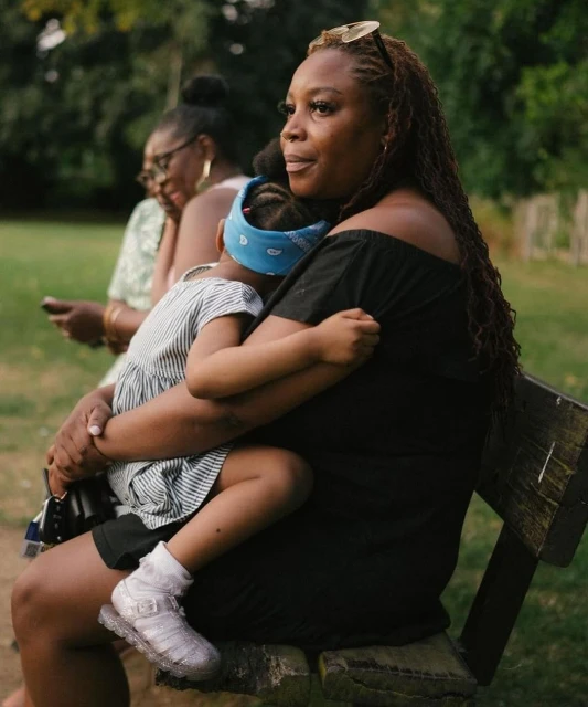 A woman in a black dress sits on a park bench holding a sleeping child with another woman sitting on the bench in the background.