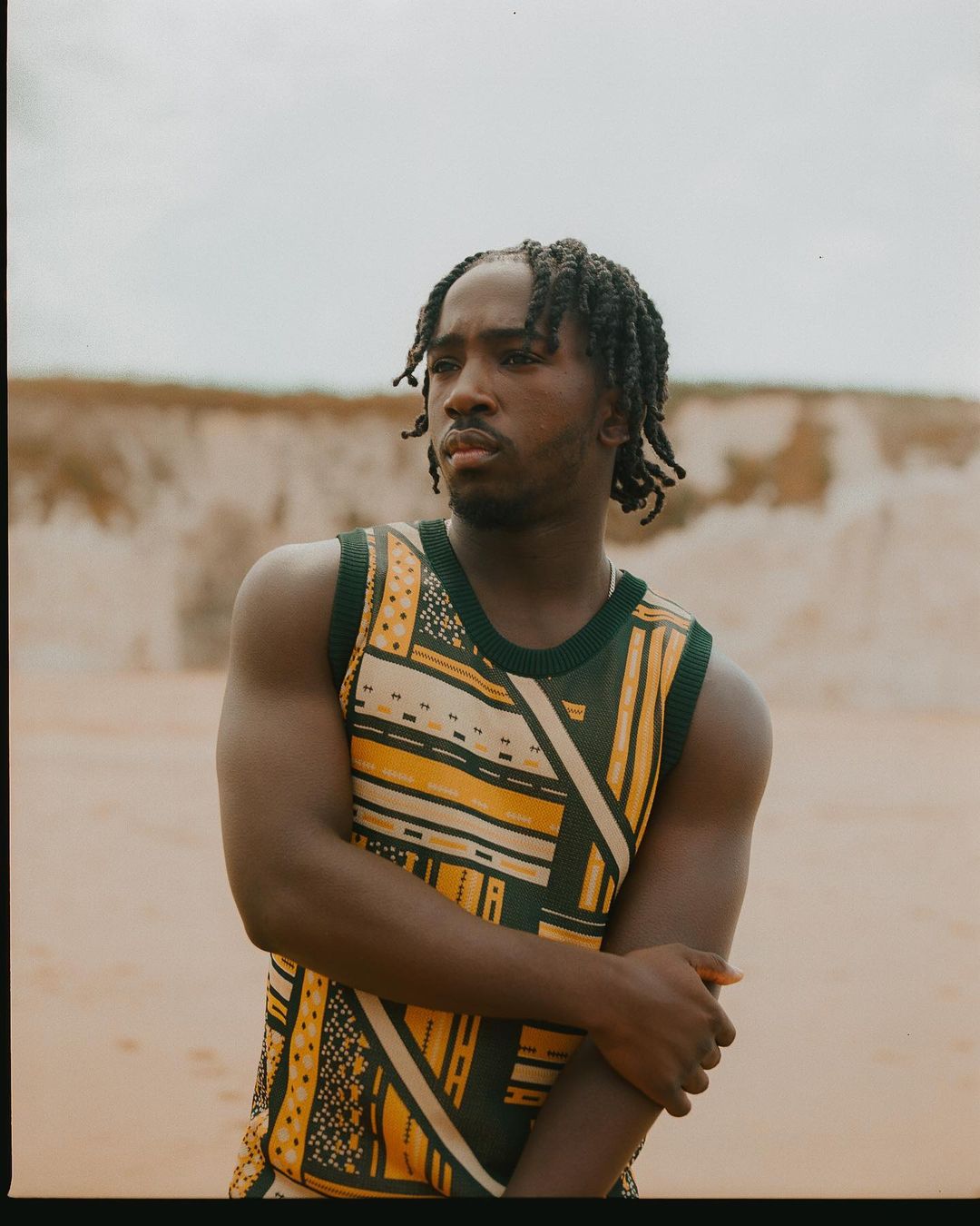 Young adult male in a patterned tank top poses in a sandy environment with a blurred background. He has dark skin and dreadlocks.