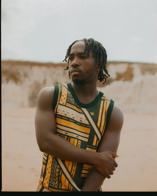 Young adult male in a patterned tank top poses in a sandy environment with a blurred background. He has dark skin and dreadlocks.