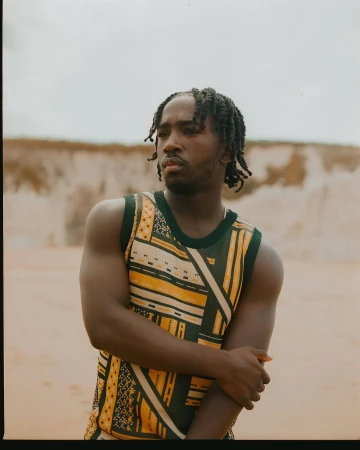 Young adult male in a patterned tank top poses in a sandy environment with a blurred background. He has dark skin and dreadlocks.