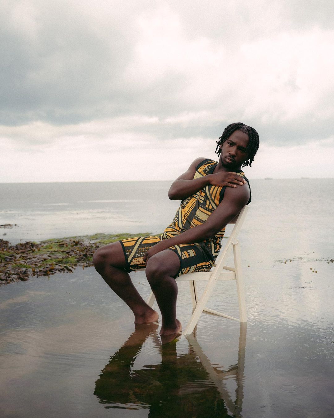 Young adult man sits on a white chair on a flooded bank near the ocean with cloudy skies. He is wearing a patterned sleeveless outfit.