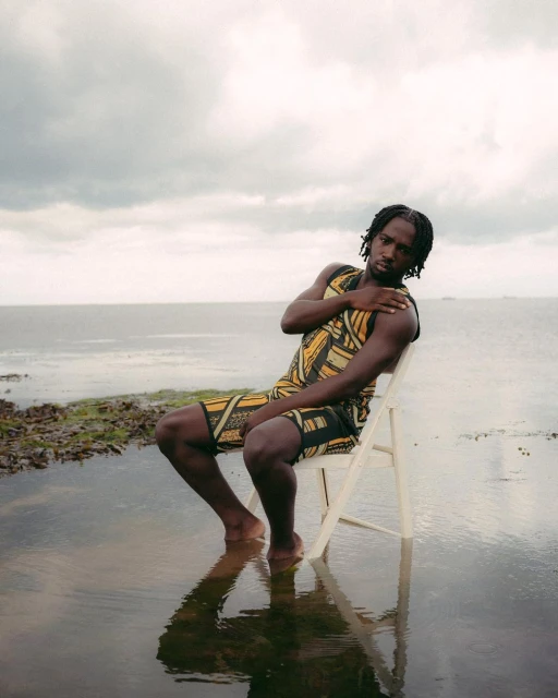 Young adult man sits on a white chair on a flooded bank near the ocean with cloudy skies. He is wearing a patterned sleeveless outfit.