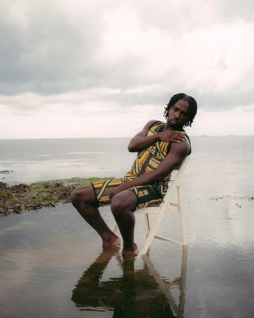 Young adult man sits on a white chair on a flooded bank near the ocean with cloudy skies. He is wearing a patterned sleeveless outfit.