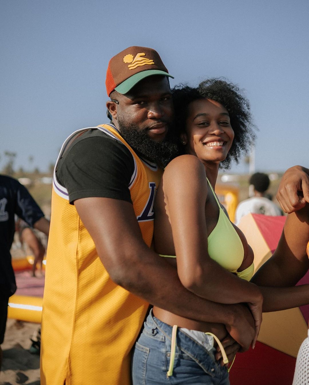 A happy couple smiles on the beach, with a man wearing a Lakers jersey embracing a woman in a yellow bikini top.
