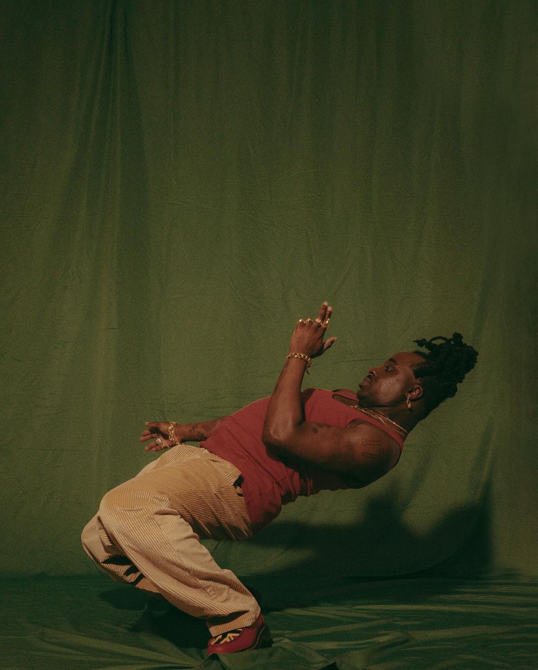 Adult male with dreadlocks in an energetic pose against a green backdrop, showcasing a red tank top and tan pants in a dynamic studio portrait.