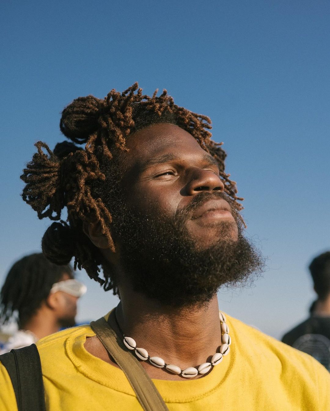 Portrait of a Black adult male with dreadlocks outdoors against a clear blue sky wearing a yellow t-shirt and a shell necklace.