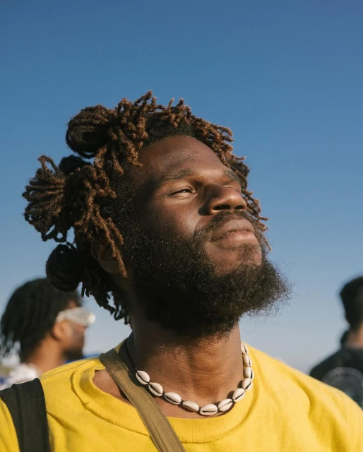 Portrait of a Black adult male with dreadlocks outdoors against a clear blue sky wearing a yellow t-shirt and a shell necklace.