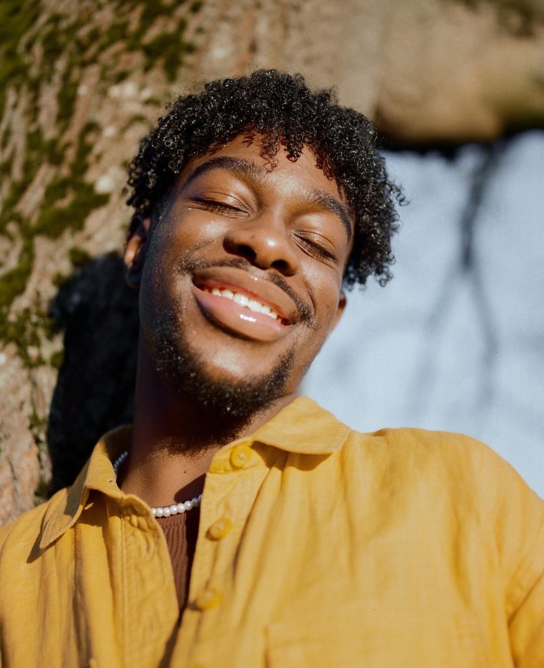 Close-up portrait of a smiling African American man with curly hair, wearing a yellow collared shirt and a pearl necklace outdoors.