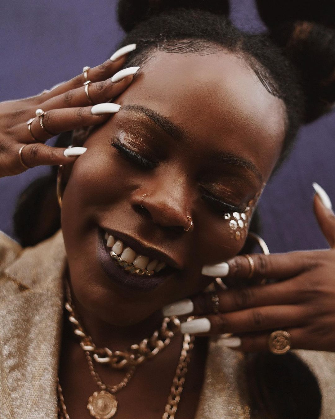 Close-up portrait of a Black woman with glitter makeup and gold teeth, wearing gold jewelry against a blurred purple background.