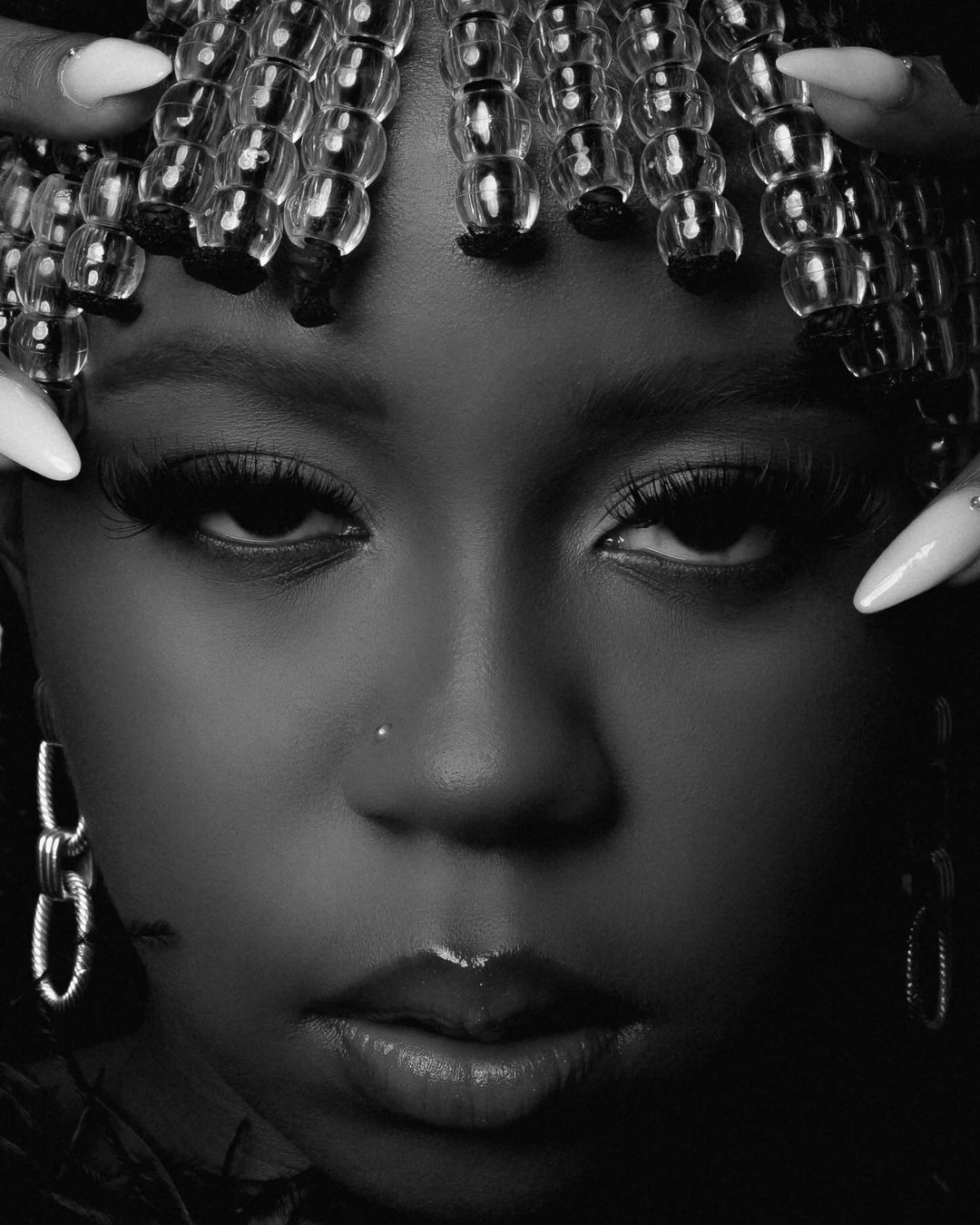Close-up portrait of a woman with dark skin and beaded hair ornaments. She has long lashes and is wearing hoop earrings in a studio shot.