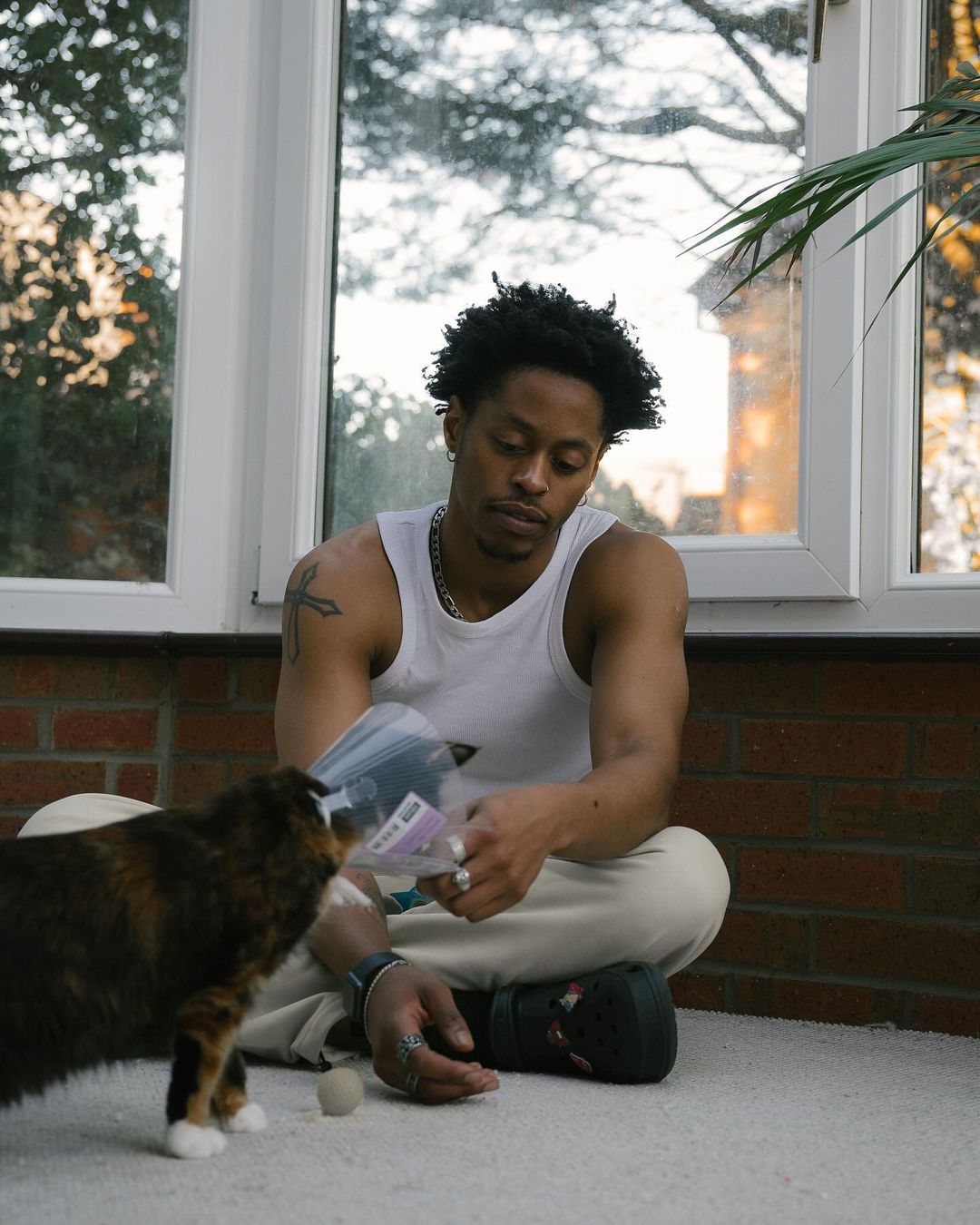 Young Black man sits indoors with his cat wearing a cone, looking down at its paws, in front of a window with plants nearby.