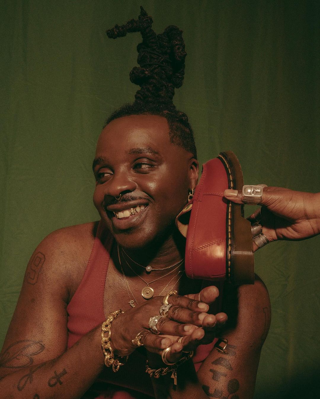Smiling African American man with a unique hairstyle holds a red leather shoe. He is wearing gold jewelry against a green backdrop.