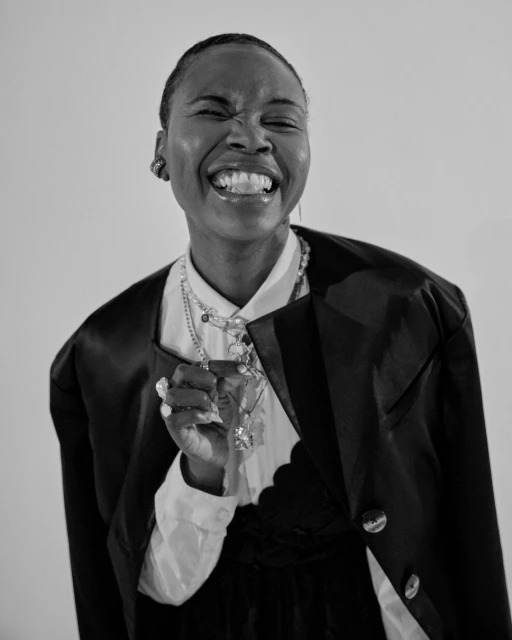 Black and white studio portrait of a joyful Black woman with natural hair, wearing a blazer, jewelry, and laughing with her mouth open.