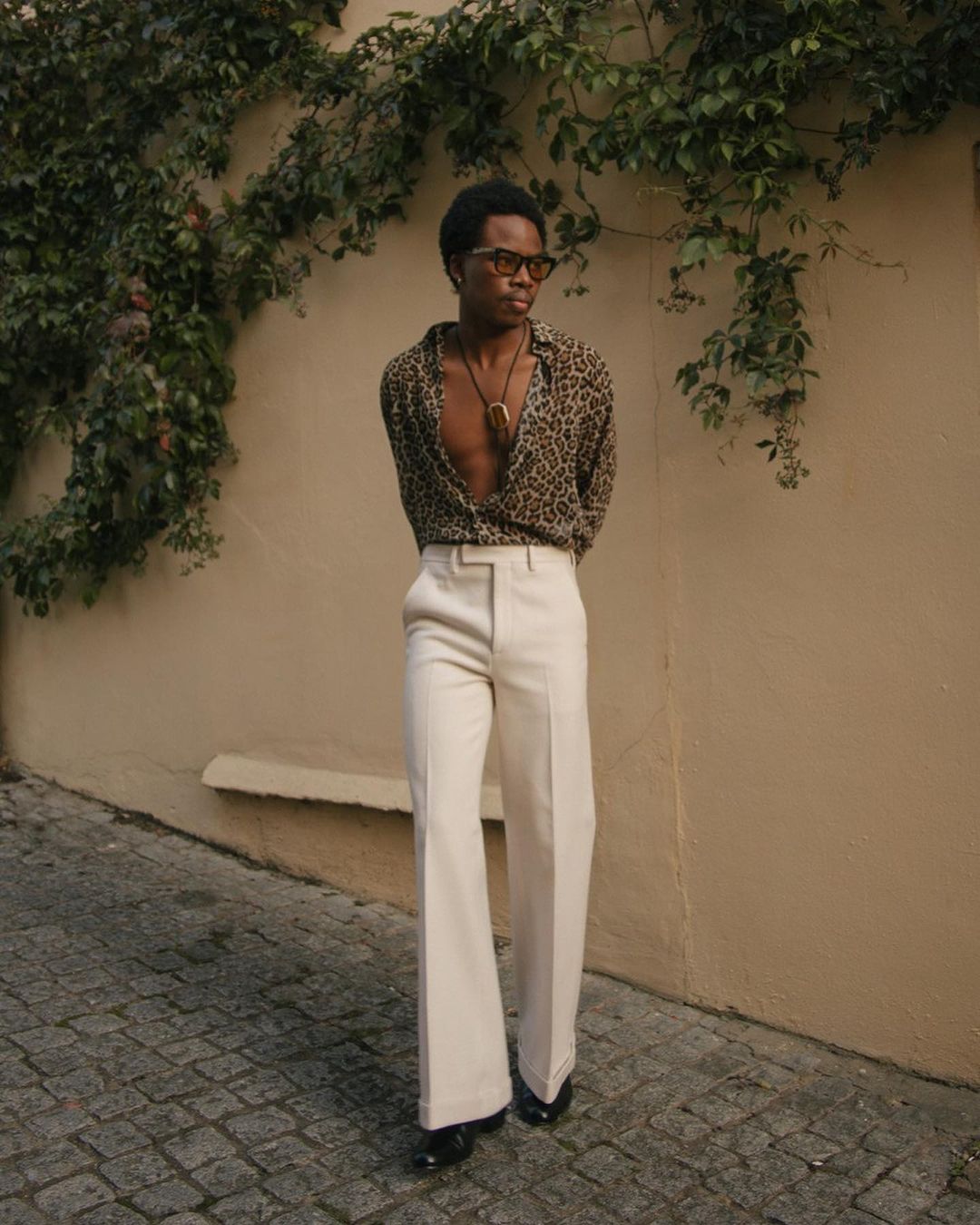 A full-length portrait of a young Black man in a patterned shirt and wide-legged pants standing on a cobblestone street next to a tan wall.
