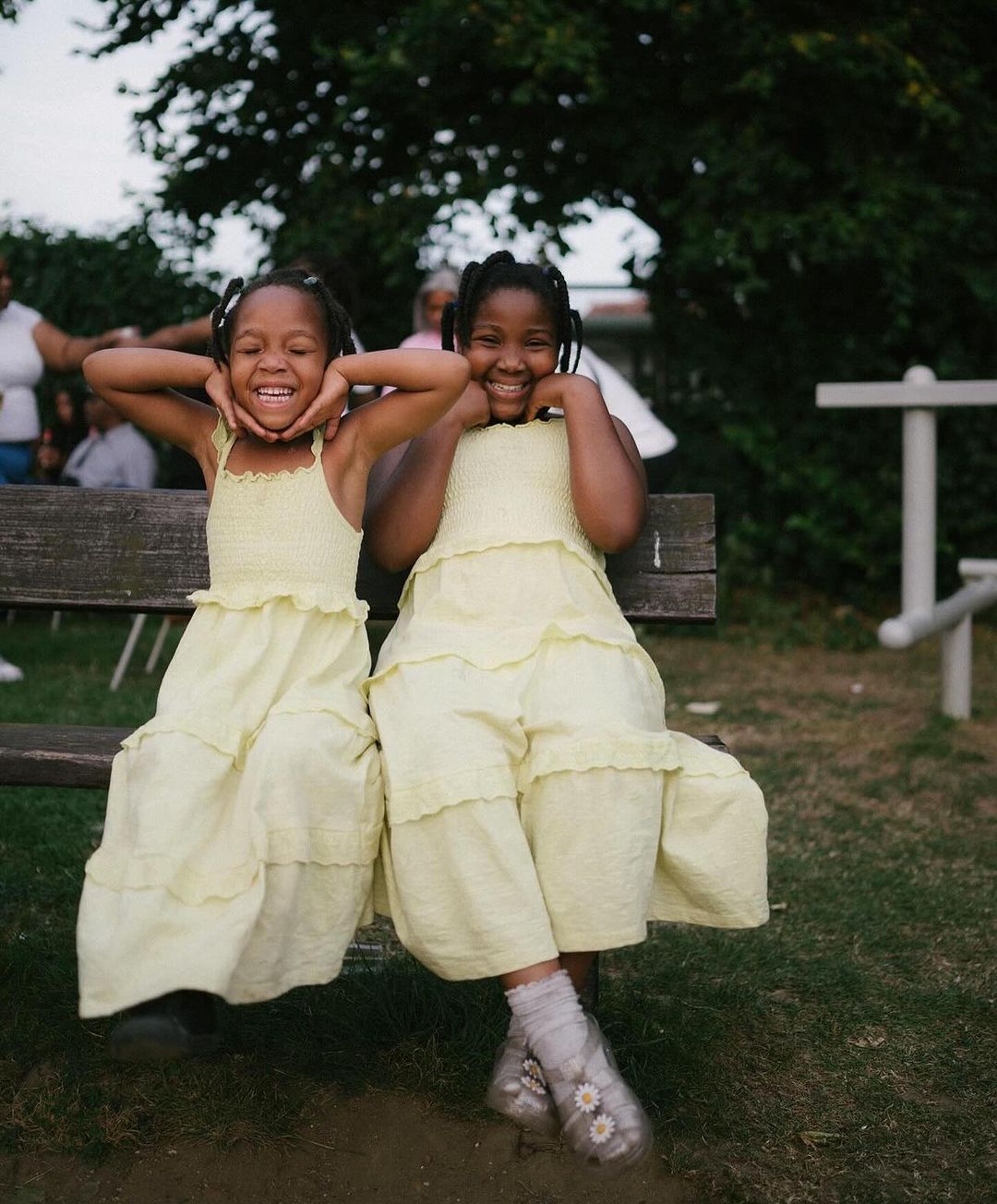 Two happy African American girls in yellow dresses smile and pose, sitting on a wooden bench in a park with lush trees in the background.