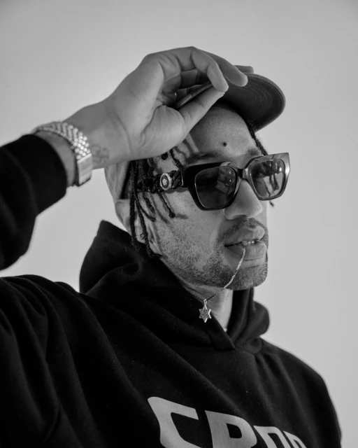 Black and white portrait of a stylish adult male wearing a cap, sunglasses, hoodie, and jewelry in a photo studio setting.
