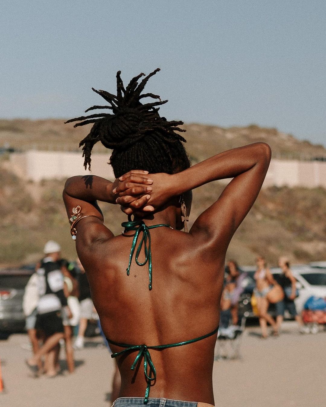 Back view of a slender woman with dark skin and dreadlocks on a beach. She wears a shiny green top and denim shorts.
